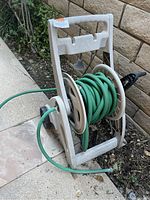 Green garden hose coiled on beige rolling hose cart with handle and wheels, placed outdoors on ground near stone wall.