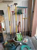 Photo showing three rakes, a garden hoe, various garden tools, garden hose, watering can, and bags of plant food on the floor.