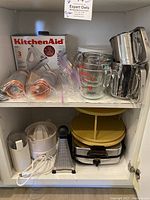 Kitchen shelf with assortment including KitchenAid Classic Mixer in box, glass measuring cups, metal sifters, decorating bags, waffle maker, cheese grater, and juicer.