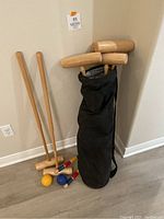 Six wooden croquet mallets leaning against a wall along with six colored balls (yellow, blue, red) and a black carrying bag containing some mallets.