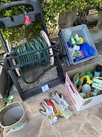 Full lot overview showing garden hose on roll-up caddy, two boxes of gardening supplies, spreader, and gloves