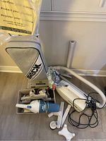 Photo showing Oreck XL upright vacuum with fabric bag, handheld Vacmaster Dustbuster vacuum, multiple white vacuum hoses and attachments including power cord, connectors, and a cross-shaped vacuum accessory base.