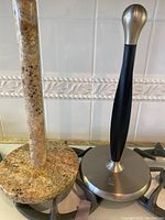 Two paper towel holders on kitchen stove, one polished brown granite and one Umbra Buckle black and silver