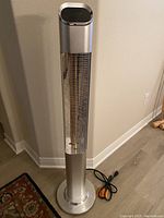 Full view of the Patio Boss tower heater fan showing metallic silver finish, cylindrical heating element, base, power cord and remote control on floor.