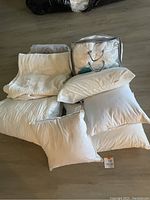 Image showing multiple white pillows, white knitted blanket, clear bag with white down comforter, and a gray blanket on the floor.