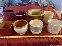Photo of 7 ceramic planter pots and a rust-colored tin pitcher arranged on a red cloth, showing variety in shapes, colors and designs of the pots and size of the pitcher.