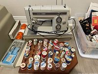 Full view of the sewing machine, bobbins, spool holder with colorful threads, box of needles, and a plastic container with additional sewing notions.