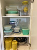 Photo inside cabinet shelf showing assortment of Tupperware containers stacked, with vintage canisters and Sun Tea glass jar with yellow lid on top shelf and bottom left