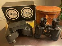 Two vintage black binoculars, one with an original black box and another with a brown leather case.