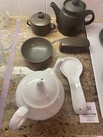 Lot showing white Apilco porcelain teapot, matching spoon holder, dark brown stoneware teapot, sugar bowl, small round dish, and butter dish lid on countertop.