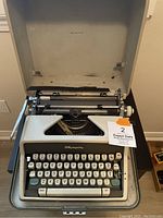 Vintage Olympia typewriter with open lid showing keyboard and platen roller. View from front with visible keys and case interior.