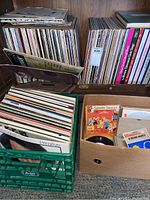Photo showing shelves and boxes filled with numerous vinyl records and albums, including a green plastic crate and cardboard box holding records and 45s in sleeves.