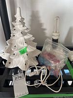 White ceramic Christmas tree next to bag of colored bulbs and plug-in base with power cord.