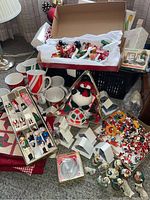 Wide shot showing assorted Christmas decorations including boxed wooden ornaments, caroller set, basket with lights, coffee mugs, dish towels, and figurines, laid out on carpet.