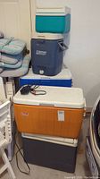 Photo showing stacked coolers including a large orange Coleman cooler with white cushioned lid, blue Coleman beverage cooler, turquoise small cooler, and gray cooler at bottom.