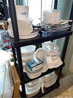 Three-tier shelf displaying stacks of CorningWare Cornflower Blue dishes with lids, plastic covers, handles and trays