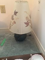 Full photo of the ceramic based lamp showing the floral shade and lamp base on carpeted floor near wall outlet.