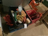 Multiple extension cords, shop lights, sponges, tape, rope in black and red crates on basement floor