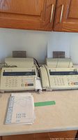Photo showing two beige Brother IntelliFAX 770 fax machines side by side with telephone handsets and keypads visible, including a user manual placed in front.