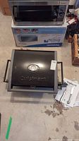 Black Cuisinart portable tabletop gas grill with box and microwave in background, two utensils and manual on side.
