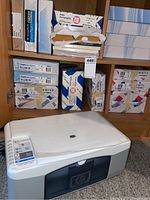 Full view of HP Deskjet F340 printer on floor with wooden shelving behind holding multiple unopened packages of bond paper.