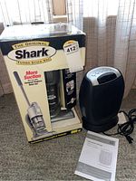 Shark Turbo Stick Vacuum in box standing next to a digital fan heater and instruction manual on carpet