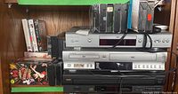 Shelf with stacked electronics including Toshiba DVD/VHS player, RCA 5-disc CD changer, and silver digital DVD player with multiple VHS movie tapes surrounding them.