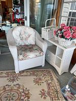 White wicker armchair with floral cushions, a white wicker side table with shelves, white wicker planter, and waste basket on display in a sunroom area.