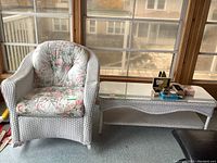View of white painted wicker rocking chair with floral seat cushion and matching wicker coffee table with glass top near window.