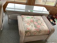 Painted wicker coffee table with glass top and painted wicker ottoman with floral cushion in front of a window with wood paneling.