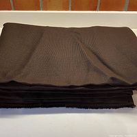 Folded chocolate brown linen upholstery fabric stack on white surface, showing thickness and texture in a wide shot.