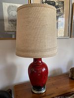 Full view of the lamp showing the red ceramic base and burlap lampshade against a neutral wall with framed artwork behind.