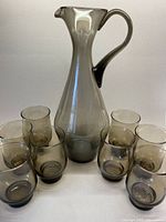 Front view of the tall smokey grey/brown glass decanter with 8 matching tulip-shaped tumblers arranged around its base on a plain background.