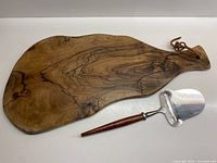 Top view of olive tree wood cheeseboard with cheese utensil placed beside it showing wood grain and handle with hanging strap.