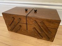 Front view of closed wooden rectangular box with two round wooden knobs on top and accordion arms on front sides.