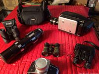 Wide shot showing the cameras, three pairs of binoculars including a large black telephoto lens and multiple carrying cases arranged on a red quilt.