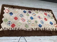 Top-down view of the rectangular rug showing the all-over design of colorful shapes in beige field with brown border, laid on wooden floor.
