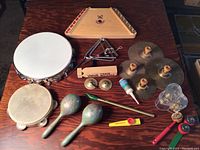 Overview of the various musical instruments on a wooden table, showing tambourines, maracas, train whistle, triangle, cymbals, kazoo, and a stringed instrument.