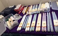 Photo showing many pairs of long beeswax taper candles in labels with a bee and honeycomb design, several colors visible, white votive candles and pillar candles in plastic containers on purple cloth.