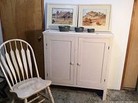 Photo shows off white wooden cabinet with two paneled doors, off white wooden chair, two framed watercolor paintings and four gray stoneware bowls on top of cabinet.