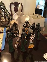 White ceramic elephant figurine standing behind six painted resin African woman figurines in various colors, displayed on wooden table.