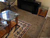 Two matching glass top end tables on carpet, showing the glass tops and wooden bases with shelves and light scratches.