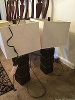 Pair of two table lamps with dark ribbed rectangular bases and white square fabric shades placed side by side on floor near a chair. One lamp cord is visible and plugged in equipment is in background.