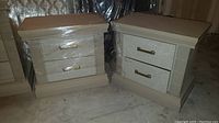 Pair of matching nightstands, boxy design, light wood finish, two drawers with brass handles, one wrapped in plastic