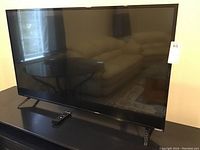 Front view of Vizio 50-inch TV on stand with remote in front on table, screen reflecting room light and furniture.