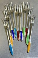 Ten sterling silver cocktail forks shown together, displaying colorful enamel handles in blue, yellow, red, green, and turquoise.