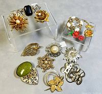 Set of 12 assorted vintage costume brooches arranged on two clear acrylic risers. Variety of floral and decorative designs primarily in gold-tone, silver-tone, and enamel with faux pearls and stones.