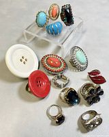 Fifteen assorted costume rings displayed on white surface with clear riser. Includes button style rings, large colorful faux gemstone rings, floral motif ring, and novelty lip ring.