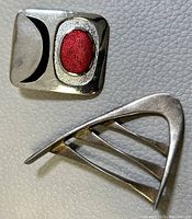 Top view of two brooches on leather: a rectangular silver enamel brooch with red and black design, and a silver modernist brooch with openwork lines.