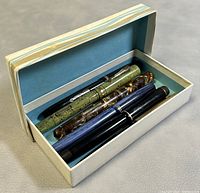 Six vintage fountain pens arranged in a light blue box, showing varying colors and patterns including green marbled, brown marbled, blue striped, and black.